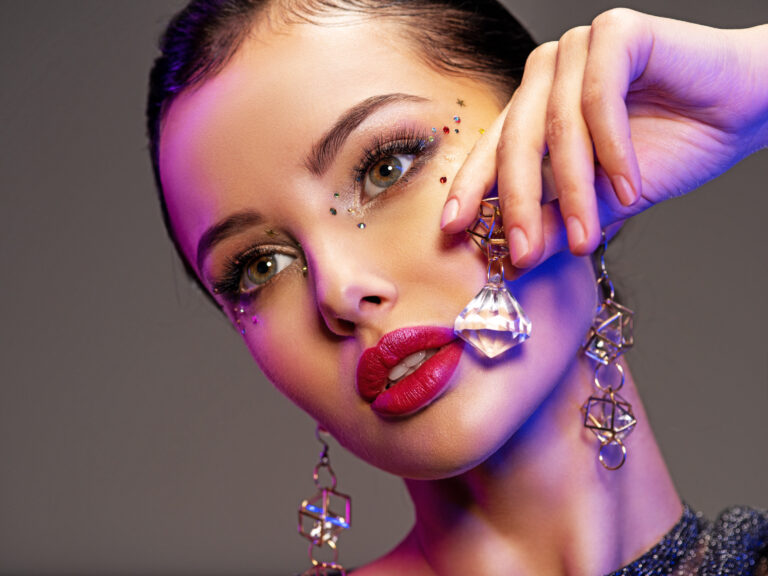 Portrait of beautiful young woman with bright makeup. Shiny sequins on face. Beautiful brunette with fashion jewelry accessories. Face of a girl is highlighted in vivid lights. Girl with earrings.