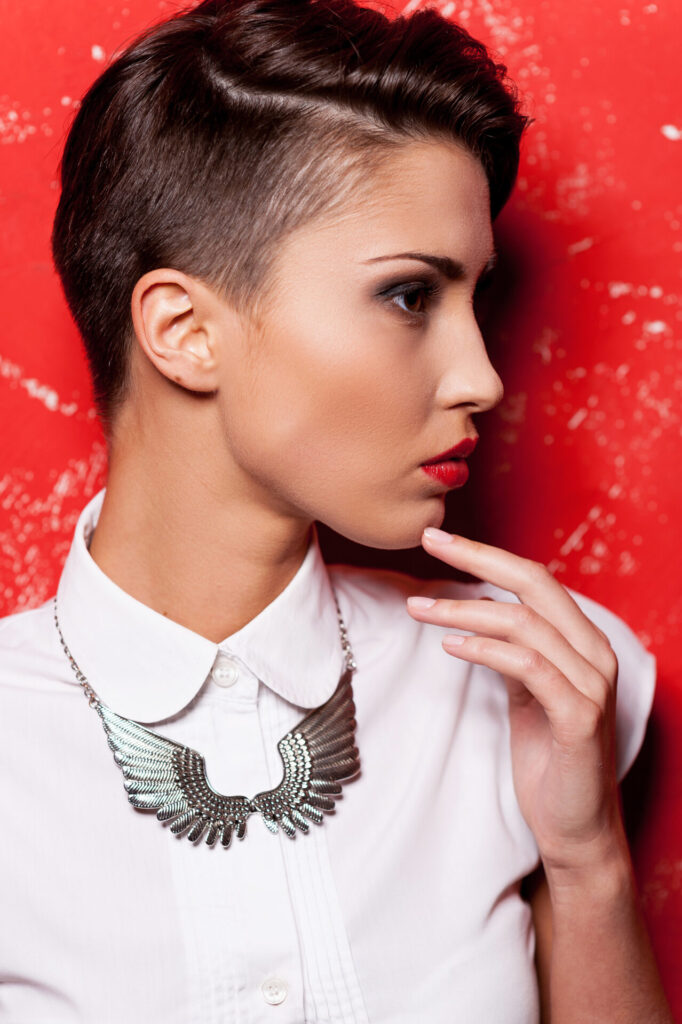 Beauty with stylish hairstyle. Side view of beautiful young short hair woman in white shirt holding hand on chin while standing against red background