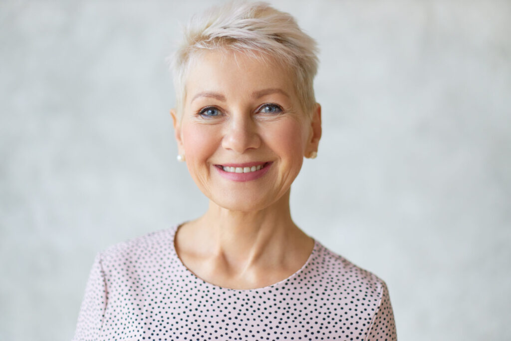 Close up image of good looking beautiful mature blonde female with blue eyes, elegant make upand pixie hairstyle smiling at camera, having confident happy facial expression, being in good mood