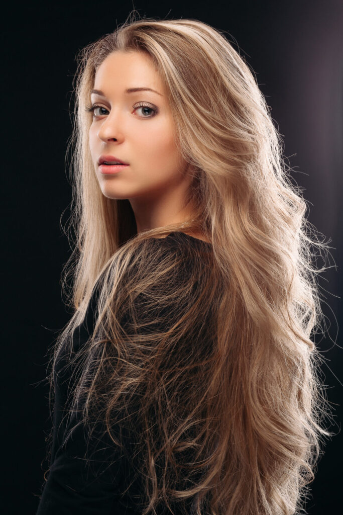 View over shoulder of beautiful and sexy blonde girl posing in studio on grey background and looking at camera. Beautiful and serious model wearing black with shiny long hair, perfect makeup.