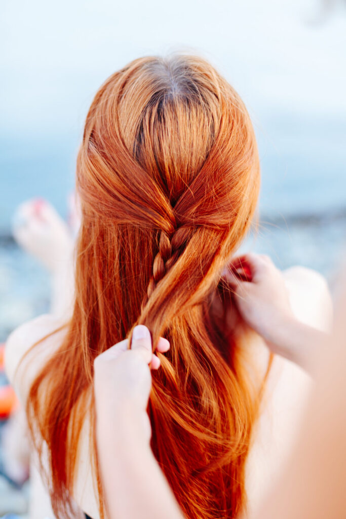 Anonymous woman standing behind redhead girl and braiding her hair on beach in sunlight.